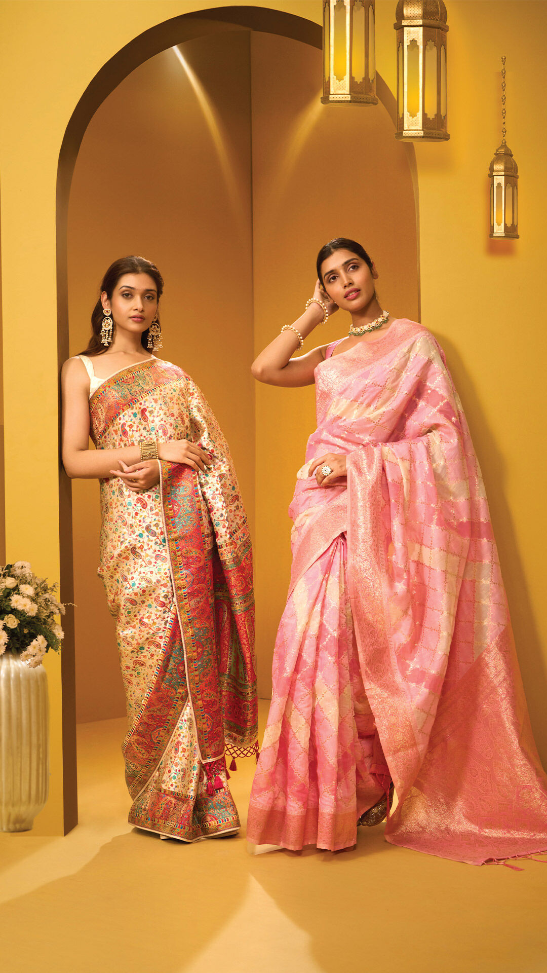 Two women in elegant wedding guest sarees — one in a vibrant multicolor brocade saree and the other in a pastel pink silk saree — posing gracefully against a mustard arched backdrop with lantern decor